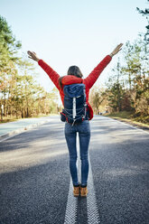 Happy woman raising arms in middle of an empty road during backpacking trip - BSZF00919