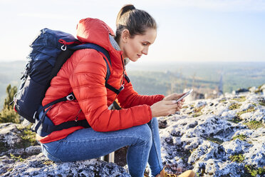 Frau beim Wandern in den Bergen, die auf einem Felsen sitzt und ihr Handy überprüft - BSZF00943