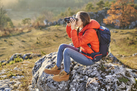 Frau auf einer Wanderung in den Bergen, auf einem Felsen sitzend und durch ein Fernglas blickend, lizenzfreies Stockfoto