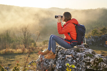 Frau auf einer Wanderung in den Bergen, auf einem Felsen sitzend und durch ein Fernglas blickend - BSZF00968