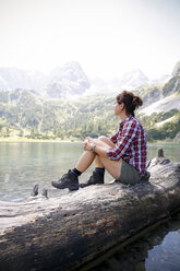 Austria, Tyrol, woman sitting on tree trunk at lake Seebensee - FKF03264