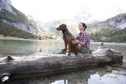 Austria, Tyrol, woman with dog sitting on tree trunk at lake Seebensee stock photo
