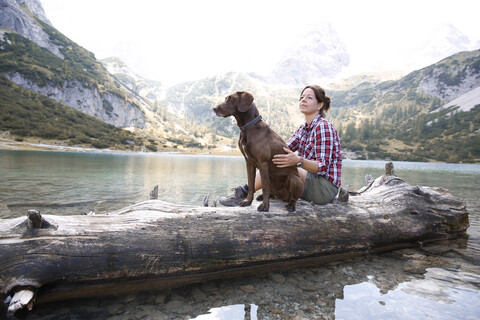 Österreich, Tirol, Frau mit Hund auf Baumstamm sitzend am Seebensee, lizenzfreies Stockfoto