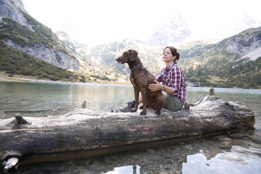 Austria, Tyrol, woman with dog sitting on tree trunk at lake Seebensee - FKF03268