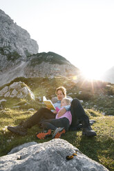 Austria, Tyrol, mother and daughter reading book in mountainscape at sunset - FKF03288