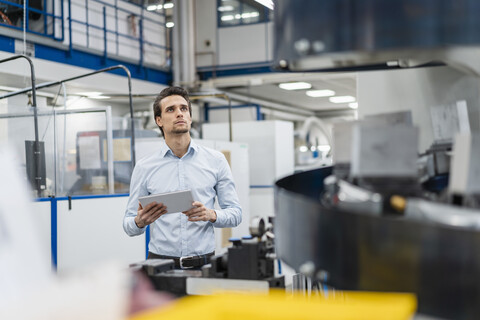Geschäftsmann hält ein Tablet und betrachtet eine Maschine in einer Fabrik, lizenzfreies Stockfoto