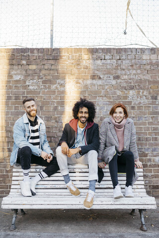Porträt von drei glücklichen Freunden, die auf einer Bank vor einer Backsteinmauer sitzen, lizenzfreies Stockfoto