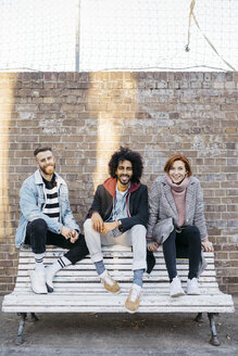 Porträt von drei glücklichen Freunden, die auf einer Bank vor einer Backsteinmauer sitzen - JRFF02633