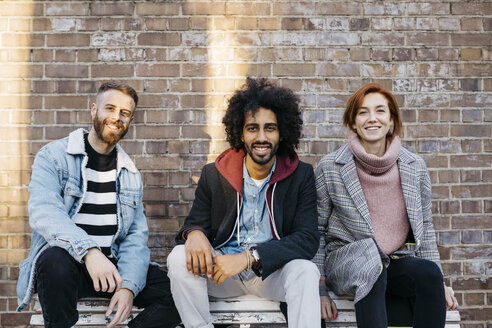 Porträt von drei glücklichen Freunden, die auf einer Bank vor einer Backsteinmauer sitzen - JRFF02634