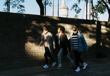 Drei glückliche Freunde gehen entlang einer schattigen Backsteinmauer - JRFF02637