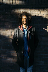 Portrait of a smiling young man standing at a brick wall in shadow - JRFF02643