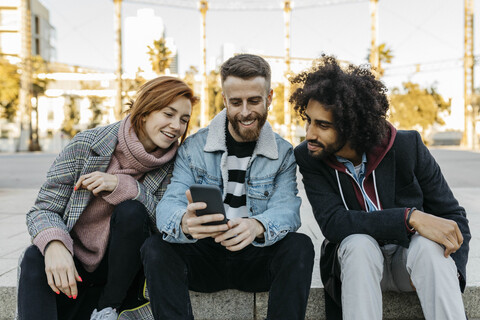 Drei glückliche Freunde sitzen im Freien und schauen auf ihr Mobiltelefon, lizenzfreies Stockfoto