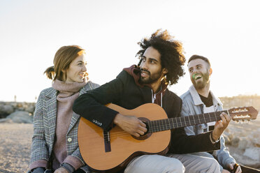 Drei glückliche Freunde mit Gitarre sitzen bei Sonnenuntergang im Freien - JRFF02652