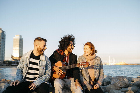 Drei glückliche Freunde mit Gitarre sitzen im Freien an der Küste Sonnenuntergang, lizenzfreies Stockfoto