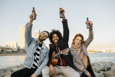 Portrait of three happy friends with guitar raising beer bottles at the coast sunset - JRFF02660