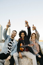 Porträt von drei glücklichen Freunden mit Gitarre, die Bierflaschen am Sonnenuntergang an der Küste hochhalten - JRFF02661