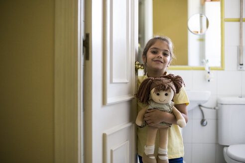 Porträt eines lächelnden kleinen Mädchens, das zu Hause im Türrahmen steht und eine Puppe hält - ABZF02230