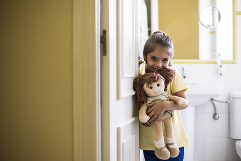 Portrait of smiling little girl standing in doorframe at home holding a doll stock photo