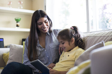 Happy mother and daughter sitting on couch at home using tablet - ABZF02233