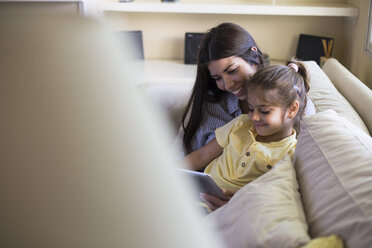 Happy mother and daughter sitting on couch at home using tablet - ABZF02234