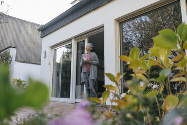 Ältere Frau mit einer Tasse Kaffee an der Terrassentür stehend - KNSF05511