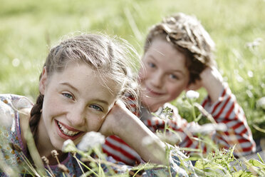 Porträt eines lächelnden Mädchens auf einer Wiese liegend mit kleinem Bruder im Hintergrund - RORF01743