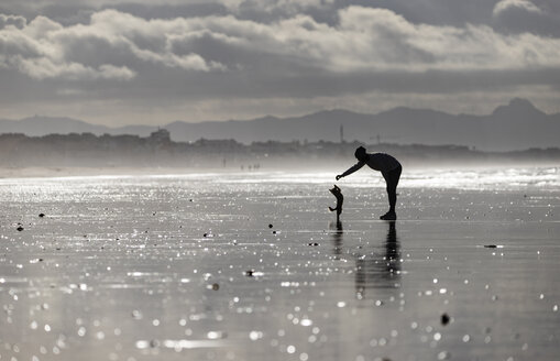 Spanien, Tarifa, Silhouette einer Frau, die mit ihrem kleinen Hund am Strand spielt - KBF00495