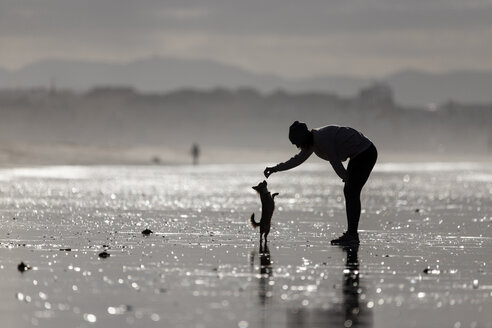 Spanien, Tarifa, Silhouette einer Frau, die mit ihrem kleinen Hund am Strand spielt - KBF00496
