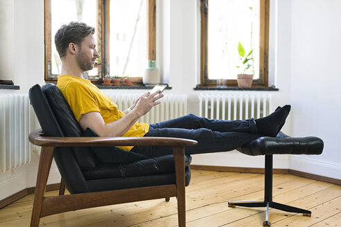 Lässiger Mann in gelbem Hemd sitzt im Lounge Chair in einer stilvollen Wohnung und schaut auf ein Tablet - SBOF01721