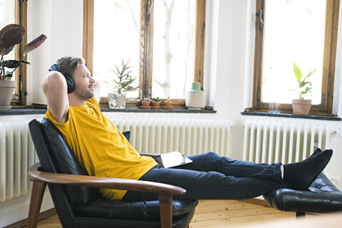 Entspannter Mann in gelbem Hemd mit Kopfhörern sitzt in Lounge Chair in stilvoller Wohnung, lizenzfreies Stockfoto