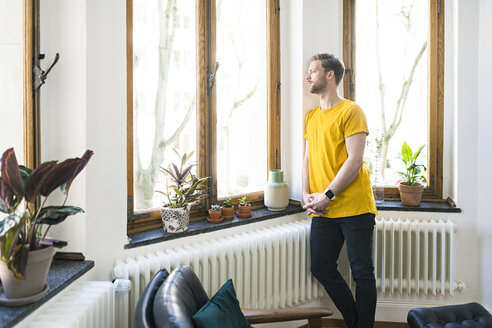 Nachdenklicher Mann in gelbem Hemd in einer stilvollen Wohnung, der aus dem Fenster schaut - SBOF01726