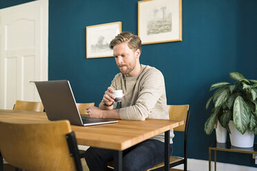 Man working on laptop on wooden table in home office drinking espresso - SBOF01728