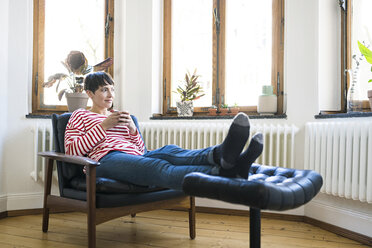 Short-haired woman relaxing in lounge chair holding coffee mug in stylish apartment - SBOF01729