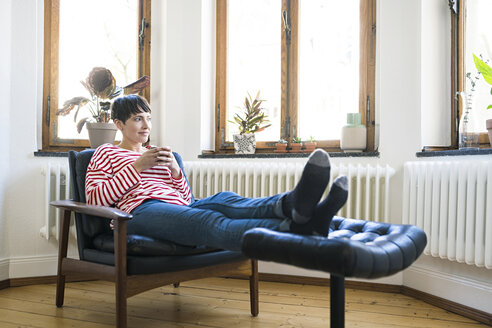 Kurzhaarige Frau entspannt sich in einem Sessel und hält einen Kaffeebecher in einer stilvollen Wohnung - SBOF01729