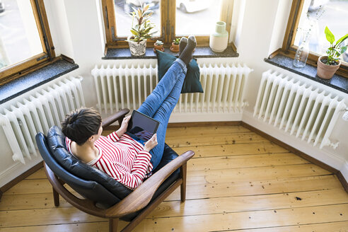 Kurzhaarige Frau entspannt sich im Sessel und hält ein Tablet in einer stilvollen Wohnung - SBOF01732