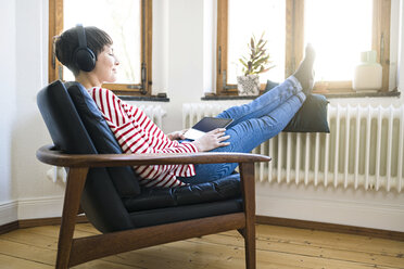 Kurzhaarige Frau mit Kopfhörern entspannt sich in einem Sessel in einer stilvollen Wohnung - SBOF01734