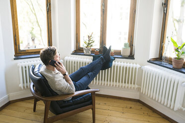 Lässiger Mann sitzt im Lounge Chair in einer stilvollen Wohnung und telefoniert - SBOF01738