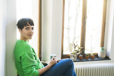 Porträt einer kurzhaarigen Frau mit grünem Pullover, die mit einem Kaffeebecher am Fenster einer stilvollen Wohnung sitzt - SBOF01741