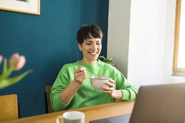 Happy short-haired woman with green pullover working with laptop in home office eating cornflakes in a bowl - SBOF01742