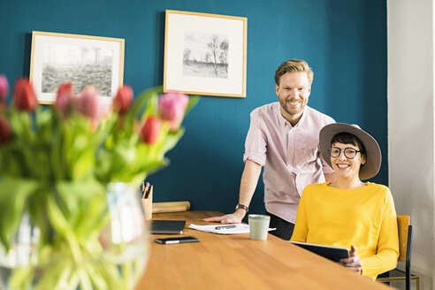 Porträt eines glücklichen Paares an einem Holztisch in ihrem Büro zu Hause mit einem Tablet - SBOF01757