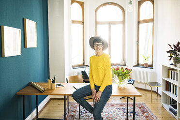 Porträt einer lächelnden Geschäftsfrau im Heimbüro, die auf einem Tisch sitzt - SBOF01761
