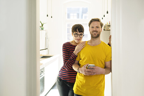 Happy couple leaning in door case in front of their kitchen stock photo