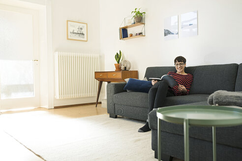 Lässige kurzhaarige Frau entspannt sich auf einer Couch im Wohnzimmer und liest eine Zeitschrift - SBOF01791
