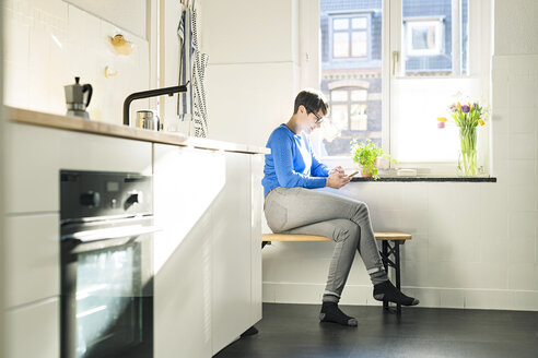 Short-haired woman sitting on bench in kitchen at the window using smartphone - SBOF01803