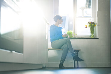 Short-haired woman sitting on bench in kitchen at the window holding coffee mug - SBOF01804