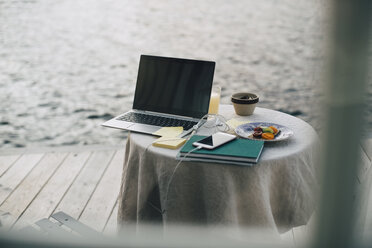 Laptop und Smartphone mit Büchern beim Frühstück auf dem Tisch durch das Fenster einer Ferienvilla gesehen - MASF11063