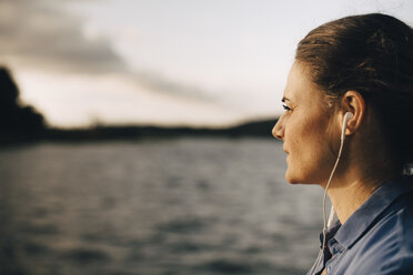 Nahaufnahme einer nachdenklichen Frau, die im Sommerurlaub am See über Kopfhörer Musik hört - MASF11078