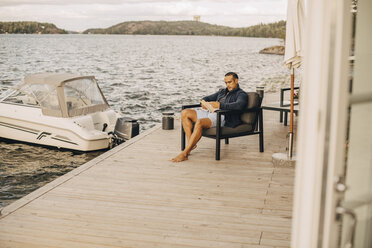 Mann in voller Länge, der ein Buch liest, während er auf der Terrasse am See sitzt - MASF11082