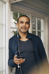 Porträt eines Mannes, der in einer Ferienvilla im Sommer über Kopfhörer auf seinem Smartphone hört - MASF11083