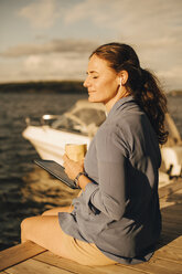 Smiling mature woman having coffee while using digital tablet at patio against lake during vacations - MASF11097